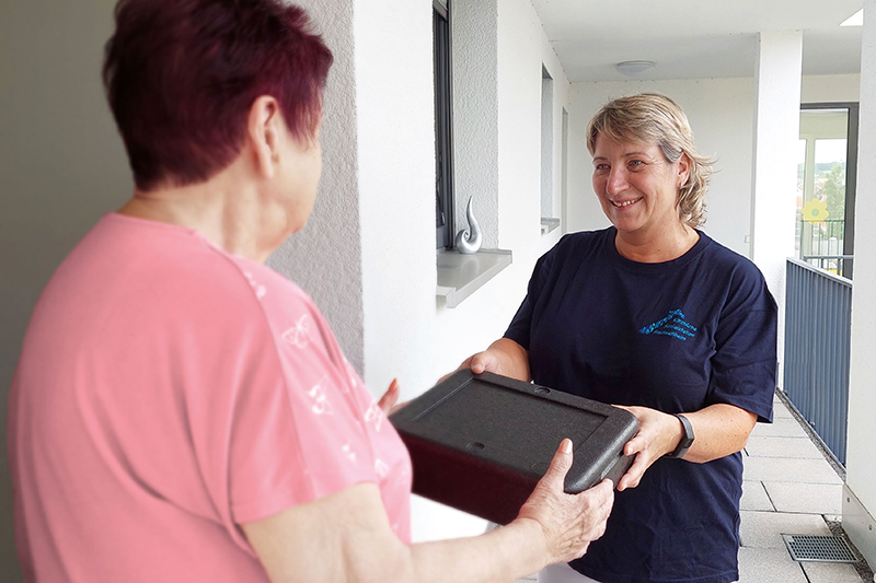Eine Mitarbeiterin übergibt eine Essensbox an eine Kundin